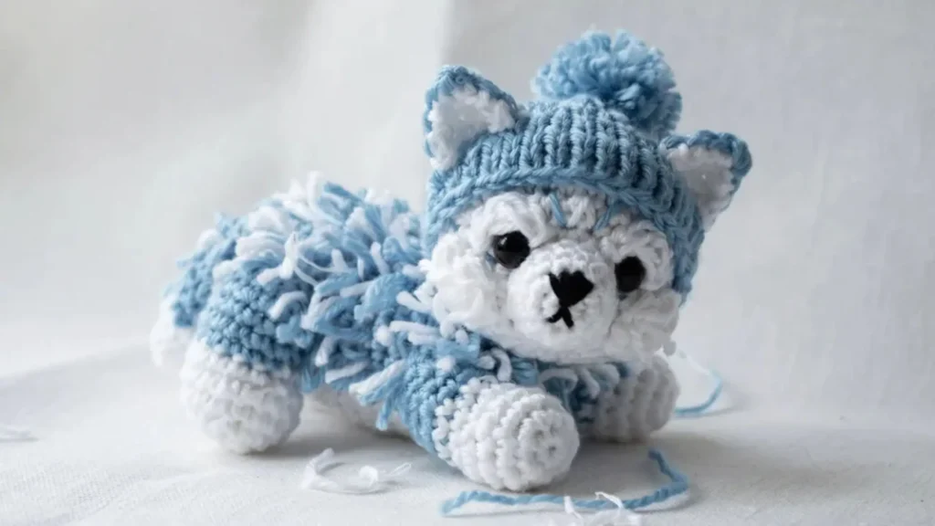 A fluffy white amigurumi crochet husky puppy toy wearing a small blue winter hat on a snowy white backdrop.