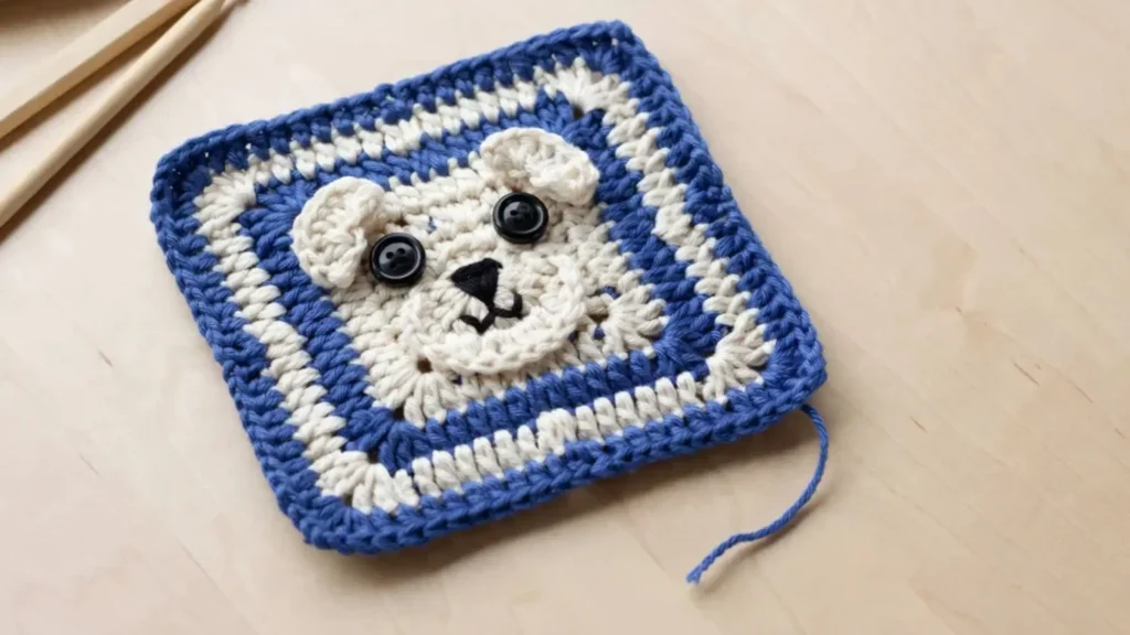  A crochet granny square featuring a stylized blue and cream puppy face design, laid flat on a wooden table.