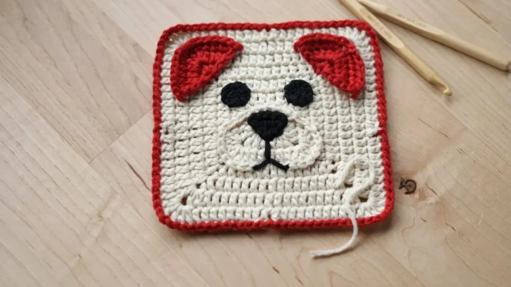 A crochet granny square featuring a stylized red and cream puppy face design, laid flat on a wooden table.