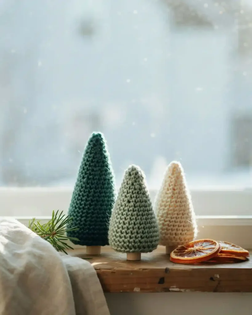 Mini crochet Christmas trees in green, sage and cream yarn displayed on a cozy winter windowsill.