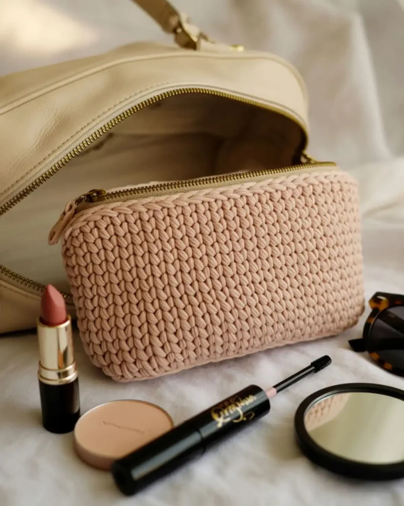 Blush pink flat crochet makeup bag with a gold zipper beside everyday cosmetics.