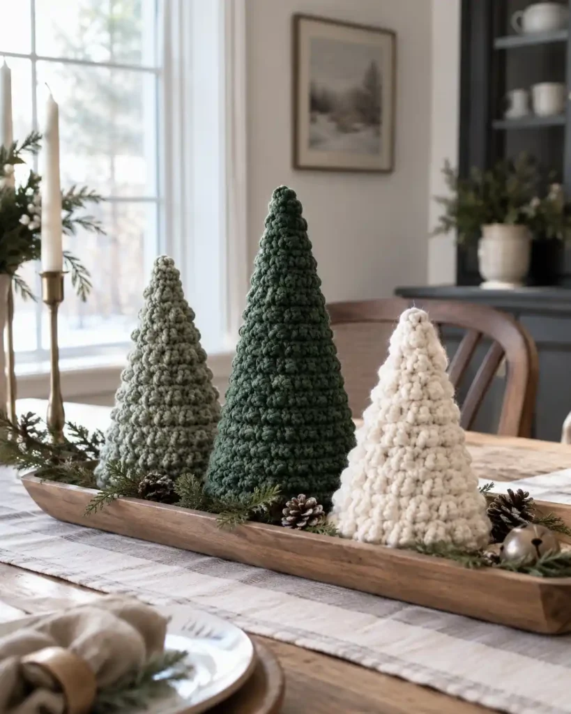 Three freestanding crochet cone Christmas trees in sage, forest green and ivory on a narrow wooden tray on a dining table.