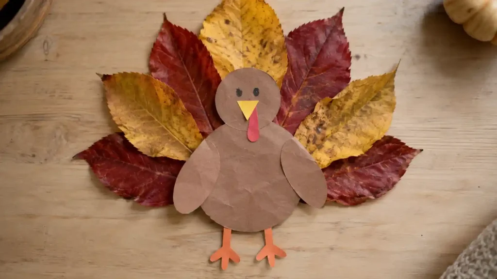 A paper turkey on cream cardstock with a fan of broad red and golden real-leaf feathers and drawn facial details.