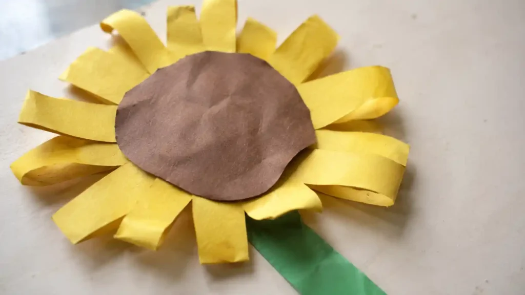 A child-made sunflower with broad yellow paper-strip petals, a large brown paper center and green paper stem.