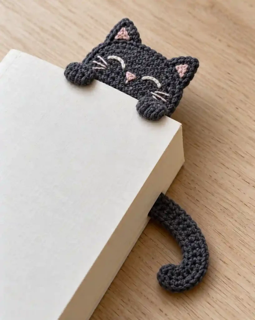 Flat charcoal crochet cat bookmark peeking out of a paperback.