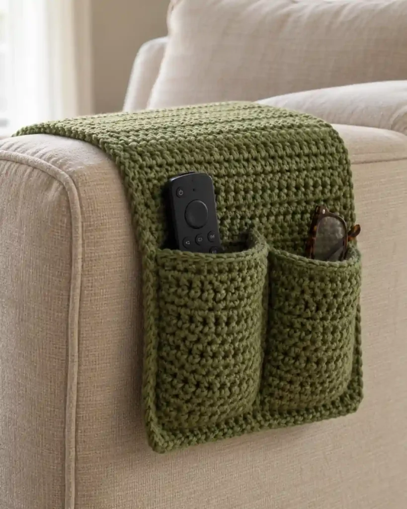 A moss green crochet caddy draped over a beige sofa arm, separate pockets holding a remote and reading glasses, side view of the fitted fabric.